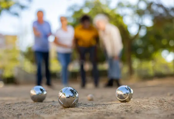 Mature casual diverse pensioners throwing petanque balls on the sand cover far away on a spring sunny day in the park