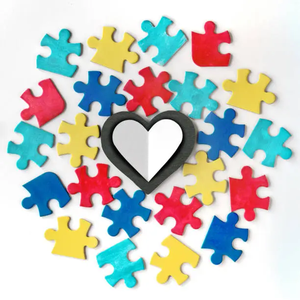 Autism campaign symbols. Colorful puzzle pieces around blackboard stone heart with copy-space for your text, lettering. Flat lay, top view, square composition on off white background.