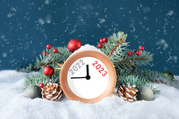 Year 2022 changes to 2023. Wood alarm clock with fir twigs, cones, red trinkets and berries. Dark green background with snow. Happy New Year 2023. stock photo