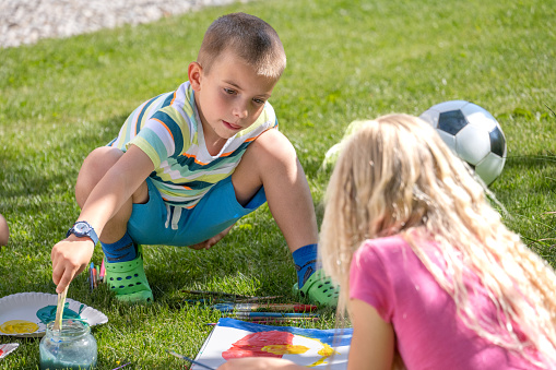 Children summer outddors activity, boy and girl painting, sitting on backyard grass. Kids leisure in nature, schoolboy and schoolgirl having fun