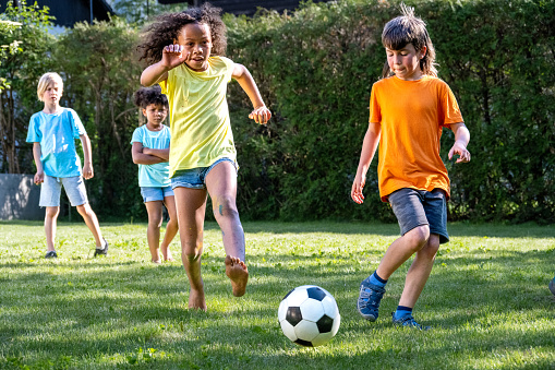 Children playing soccer outdoors, african american girl kicking football ball. Kids leisure activity in park, summer entertainment, wide shot