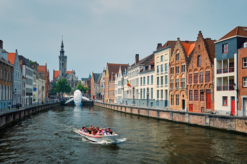 Bruges, Belgium - May 29, 2018: Tourist boat in canal between old houses with The Bruges Whale plastic installation and The Burghers' Lodge Poortersloge tower. Brugge Bruges, Belgium