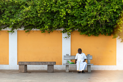 Pondicherry, India - January 2020: An Indian man sitting on a stone bench in front of a vintage yellow wall of an old building in the heritage town of Puducherry.