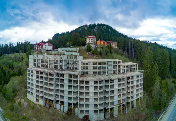 Multi-storey abandoned hotel with concrete walls stands in dense spruce forest on slope of high valley of Rhodope Mountains. Panorama, top view
