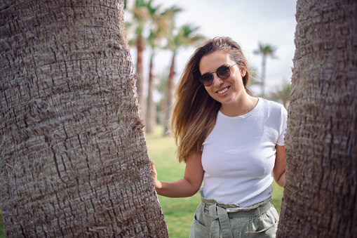 A view from the waist up of a beautiful woman standing next to a palm tree in the park.
