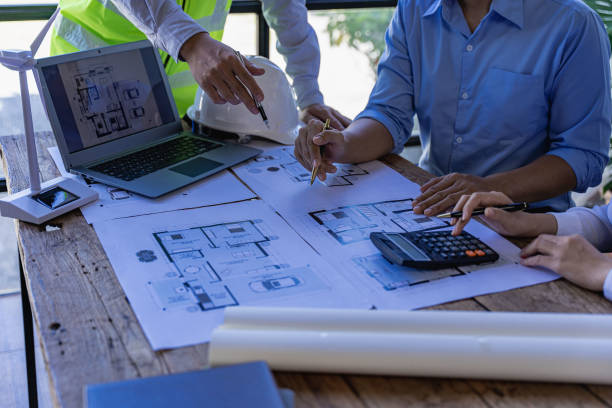 istock Architects and engineers point to buildings on blueprints and use laptops to draw house project designs in a construction concept office. 1440659190