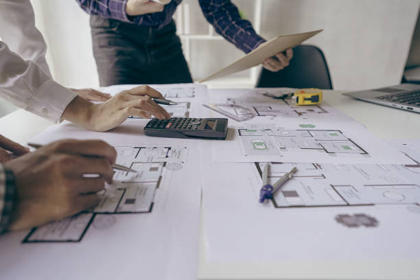 Architects and engineers point to buildings on blueprints and use laptops to draw house project designs in a construction concept office.
