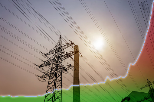 High voltage pylon and industrial chimney at sunset. A line diagram with a color gradient from green to red is shown as a symbol for the rising energy costs.