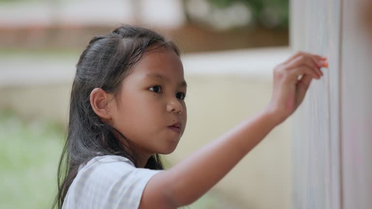 Young asian child girl writing and drawing with chalk on board at outdoor playground. Kid enjoy outdoor activity playing and learning at school playground.