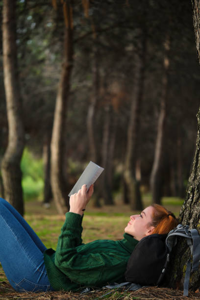 Redhead young woman reading a book lying under tree in forest.