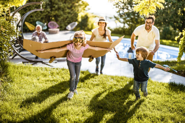 Happy little kids running like airplanes while being chased by their parents in the backyard. There are people in the background.