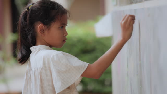 Young asian child girl writing and drawing with chalk on board at outdoor playground. Kid enjoy outdoor activity playing and learning at school playground.