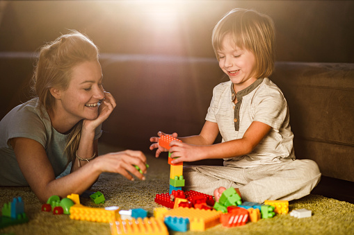 Happy mother and her small son enjoying while playing with toy blocks on carpet in the living room.