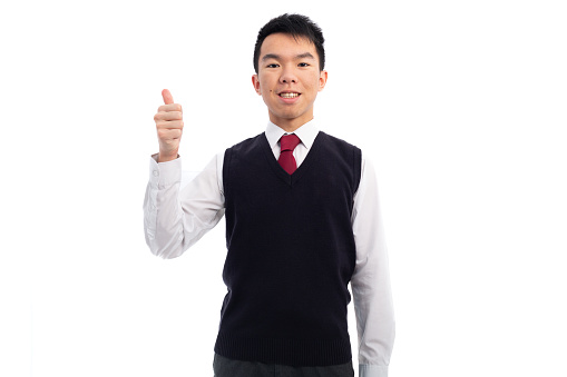Medium shot of a young teen Asian boy in the studio with broad flash lighting. He is gesturing thumbs up and grinning at the camera. He is wearing the school uniform of a white shirt with a white tie and a black sweater vest. The background is white.