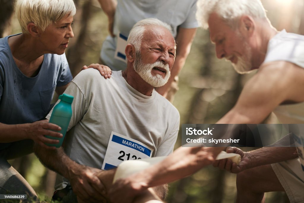 Ugh, my knee hurts so bad! Mature male marathon runner having pain in his knee while others are helping him. Active Lifestyle Stock Photo