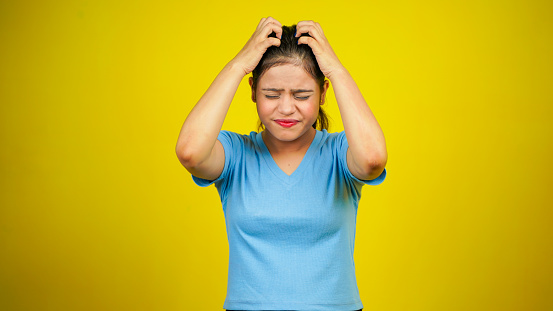 Young nervous woman scratches her head, hair itching on yellow background