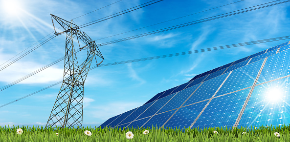 Group of solar panels and a high voltage tower on a green meadow (green grass and daisy flowers) against a blue sky with clouds and sunbeams.