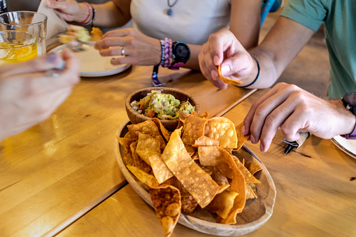 Unrecognizable people taking tortilla chips from container to eat. Anonymous friends enjoying plate of nachos with green salsa at restaurant table. Unidentified adult people eating appetizers in restaurant.