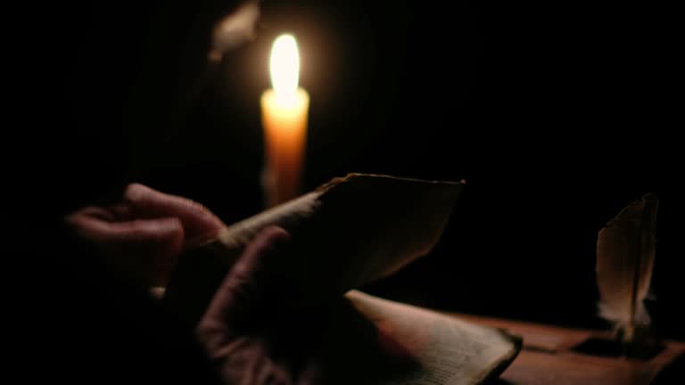 Video of old monk reading old book in dark monastic cell room under candle light.