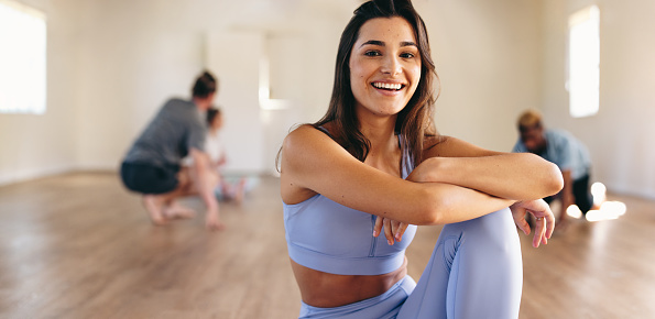 Portrait of a fit young woman smiling at the camera while sitting in a yoga studio with her class in the background. Happy young woman having a workout session in a fitness studio.