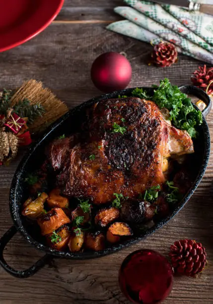 Homemade oven roasted turkey shank for christmas meal. Served in a roasting pan with vegetables on rustic and wooden table background from above with red ornaments.