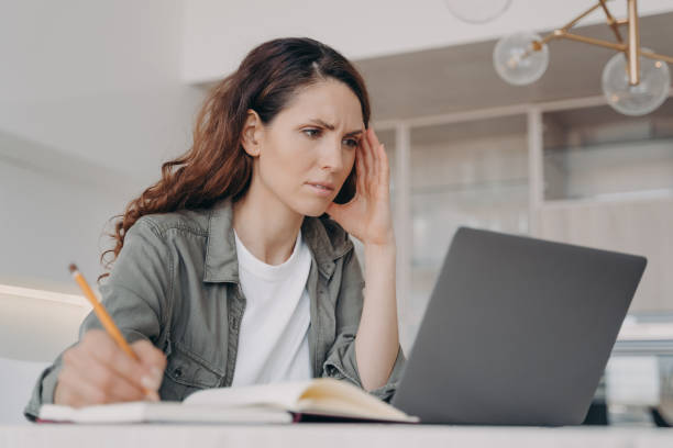 istock Frowning female student learning online at laptop, taking notes, sitting at desk at home. E-learning 1440706558