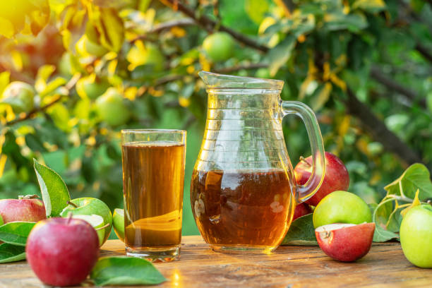 Fresh apple juice and organic apples on wooden table.  Summer orchard in the evening sun rays at the background. Fresh apple juice and organic apples on wooden table.  Summer orchard in the evening sun rays at the background. apple juice photos stock pictures, royalty-free photos & images