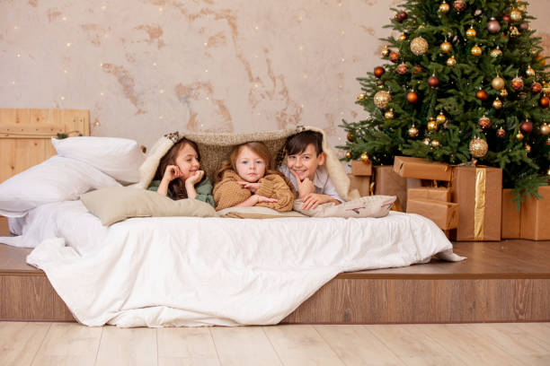 troje dzieci, dwie dziewczynki i chłopiec, szczęśliwi razem w domu, leżą na łóżku pod kocem przy udekorowanej choince - playground snow winter little girls zdjęcia i obrazy z banku zdjęć
