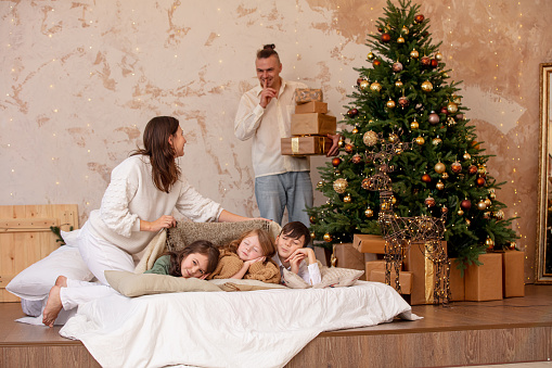 Family - father, mother, and three children, happy together at home in the bedroom, hiding gifts under the Christmas tree