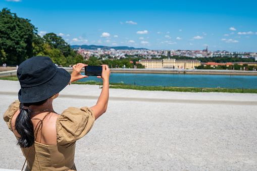 Female traveler taking a photo of panoramic view of Vienna downtown and the Schonbrunn palace from a high viewpoint on a sunny day using a smart phone. Austrian landmark view and its capital city skyline
