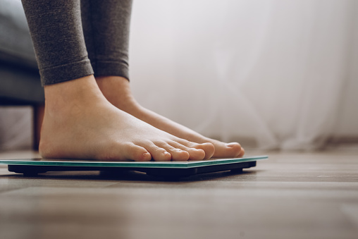 Side view of unrecognizable young woman weighing on scales at home. Female dieting checking and weight loss. Barefoot measuring body fat overweight. Control, wellness, lifestyle and diet concept