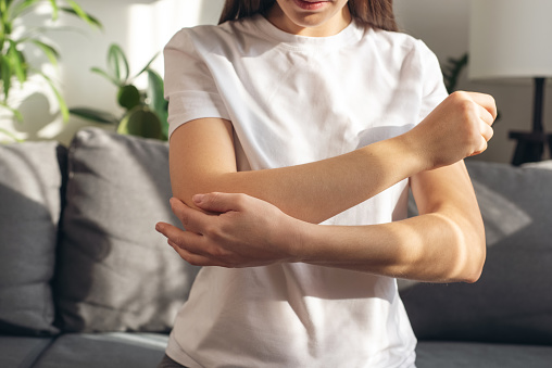 Close up of young woman with arm pain. Unhealthy caucasian girl suffering from elbow pain and massaging painful hand sitting on couch at home. Female hand holding her elbow