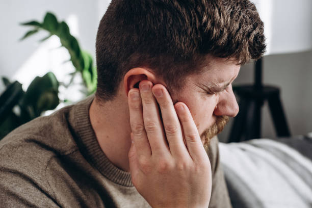 close-up of unhealthy young caucasian man 30s touching ear, suffering from sudden throbbing ear ache, looking aside. upset bearded male feeling unwell sitting on couch in living room at home - aside imagens e fotografias de stock