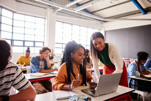 enseignant souriant parlant avec un élève à l’aide d’un ordinateur portable pendant une leçon en classe - enseignant photos et images de collection