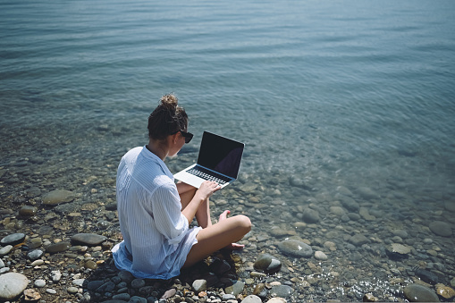 Young woman freelancer traveler wearing white shirt anywhere working online outdoors using laptop enjoying lake view. Happy female downshifter in sunglasses holding computer at sea coastline