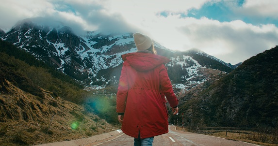 Woman in red jacket on winter travel adventure walk on empty mountain road surrounded by beautiful nature. Tourist female on hiking trip explores earth