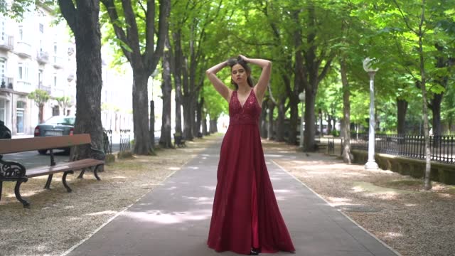 Caucasian woman with long tail red dress walking next to a park in the city, lifestyle