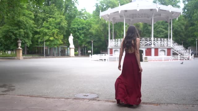 Caucasian woman in a long-tailed red dress walking next to a kiosk in the city, lifestyle
