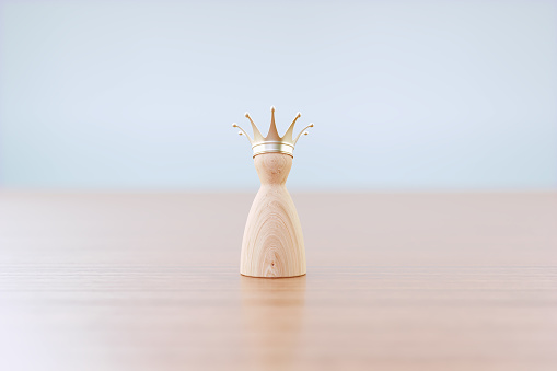 Crown wearing wood pawn standing on wood surface before defocused grey background. Horizontal composition with selective focus and copy space. Success, leadership and standing out from the crowd concept.