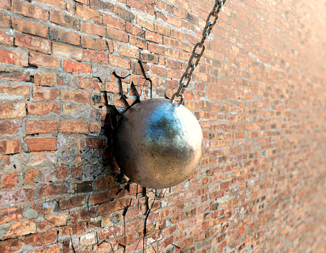 A regular metal wrecking ball attached to a chain breaking a hole in a flat brick wall surface  - 3D render