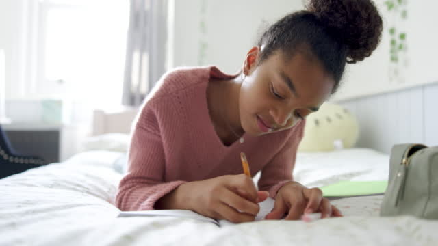 Black teenage girl lying on front on her bed doing homework while smiling