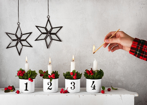 Girl hand lighting the fourth candle on a handmade advent wreath decorated with red checkerberries.\nChristmas home decoration concept. Copy space.
