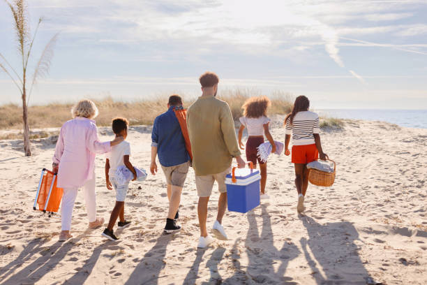 rückansicht von eine mehrgenerationenfamilie, die am strand spazieren geht und strandsachen trägt - family grandmother multi generation family nature stock-fotos und bilder