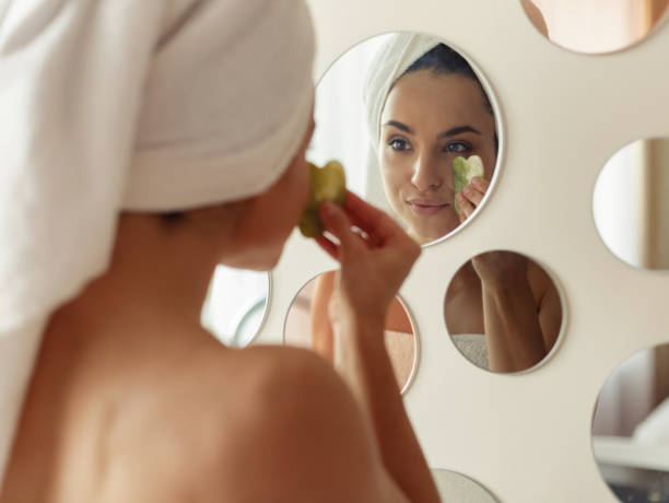 매력적인 만족 젊은 백인 여성은 괄사를 사용하여 거울을 보면서 욕실에서 얼굴 마사지기로 마사지를하는 머리에 수건을 가지고 있습니다. 집에서 피부 관리. 클로즈업 - bathtub women naked human face 뉴스 사진 이미지