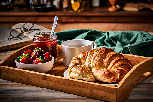 Breakfast with cup of coffee, croissants, marmalade, and fruits on wooden table in a rustic kitchen.