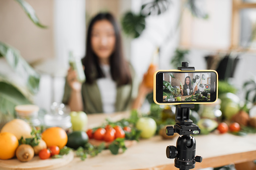 Asian woman making healthy food using phone creating video content for social media in tropical resort kitchen studio. Beautiful female using vegetables and fruits preparing smoothies