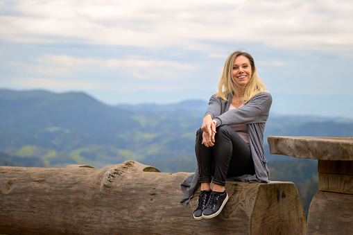 Happy laughing middle-aged blond woman sitting on a tree trunk on a mountain and cloudy sky