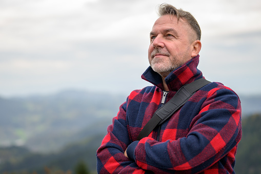 Middle aged grey-haired man with a beard and a lumberjack jacket looks into the distance on a mountain in the fog