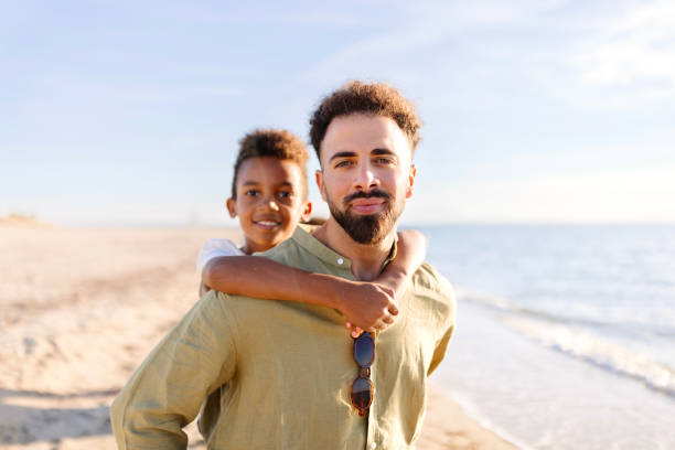 Happy father giving his little boy a piggyback ride on the beach Three generation family on a beach parent piggybacking kid father son grandfather fathers day stock pictures, royalty-free photos & images