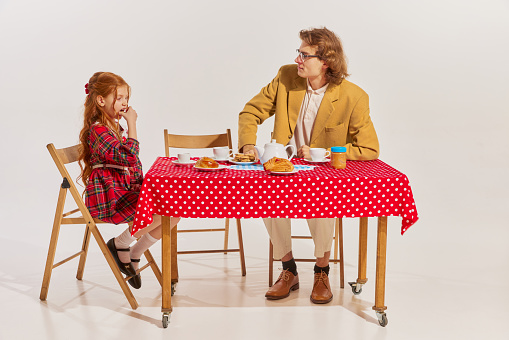 Portrait of man in jacket having breakfast with his daughter, sitting at the table isolated over grey background. Concept of beauty, retro style, fashion, elegance, 60s, 70s, family. Copy space for ad